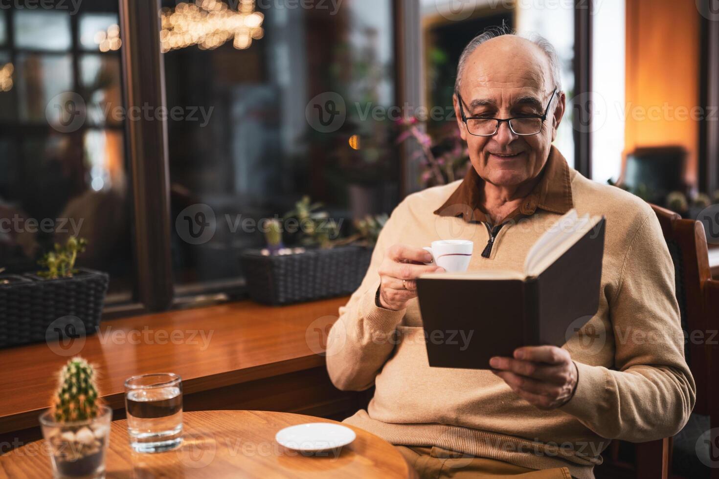 Senior man with eyeglasses reading a book and drinking coffee while sitting at the table in cafe at evening. photo