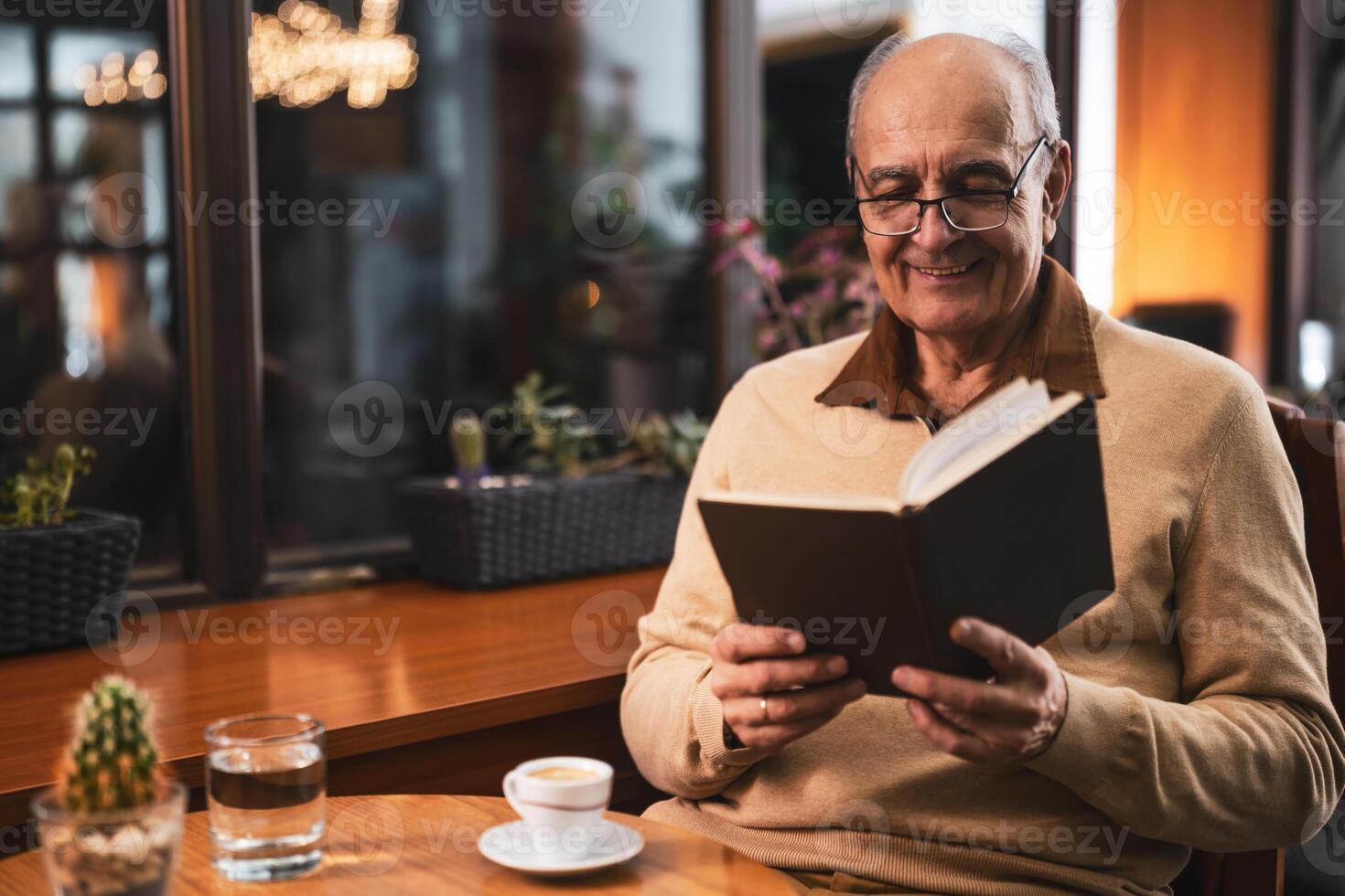 Senior man with eyeglasses in casual clothing enjoying reading a book and drinking coffee while sitting at the table in cozy cafe at evening. photo