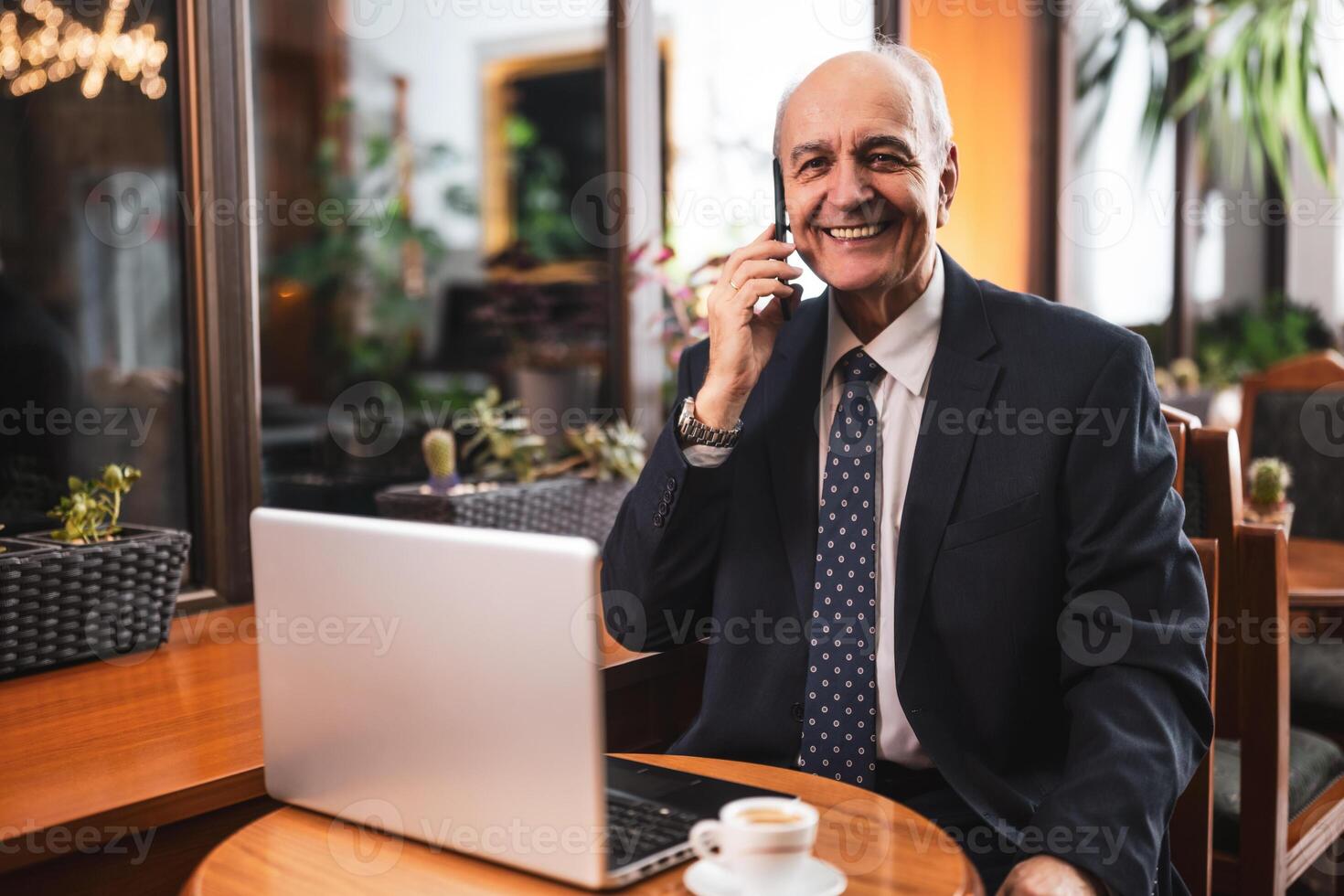 Senior businessman in elegant suit talking on the smart phone and using laptop while drinking coffee and sitting in the cozy cafeteria. photo