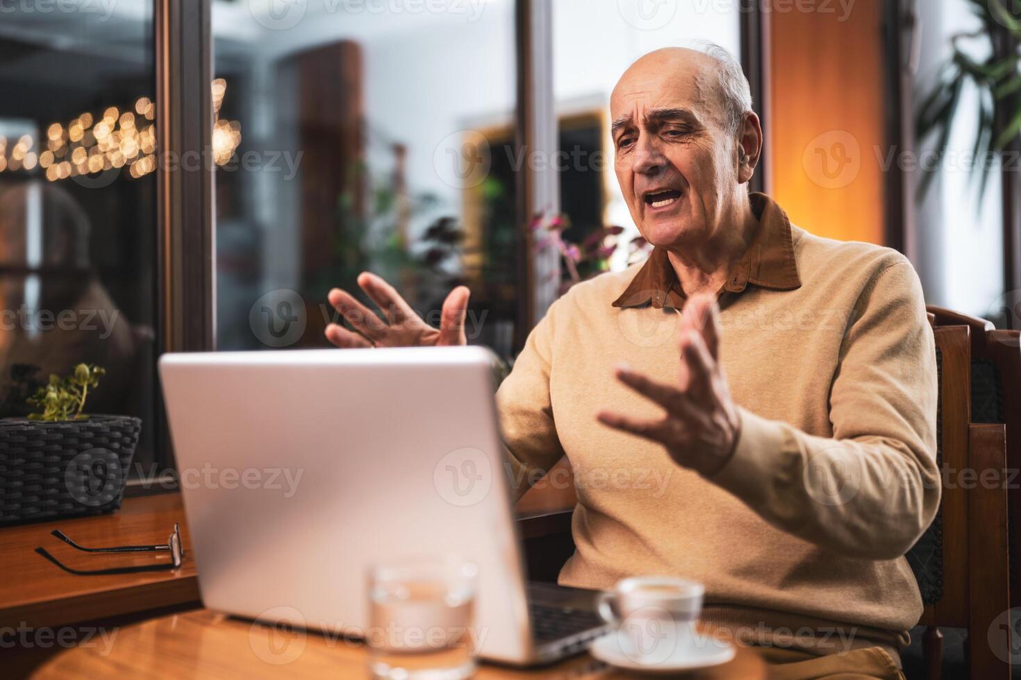 Senior man talking and gesturing while having a call on his laptop. He is using computer and drinking coffee and relaxing in the city cafe. photo