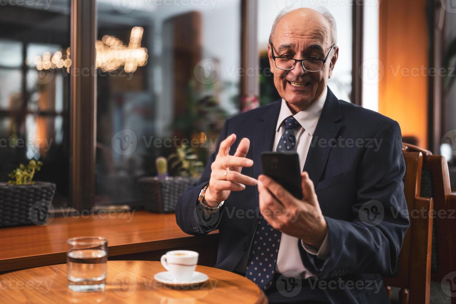 Smiling successful senior businessman with eyeglasses in elegant suit using mobile phone and drinking coffee while sitting in the cozy city cafe. photo
