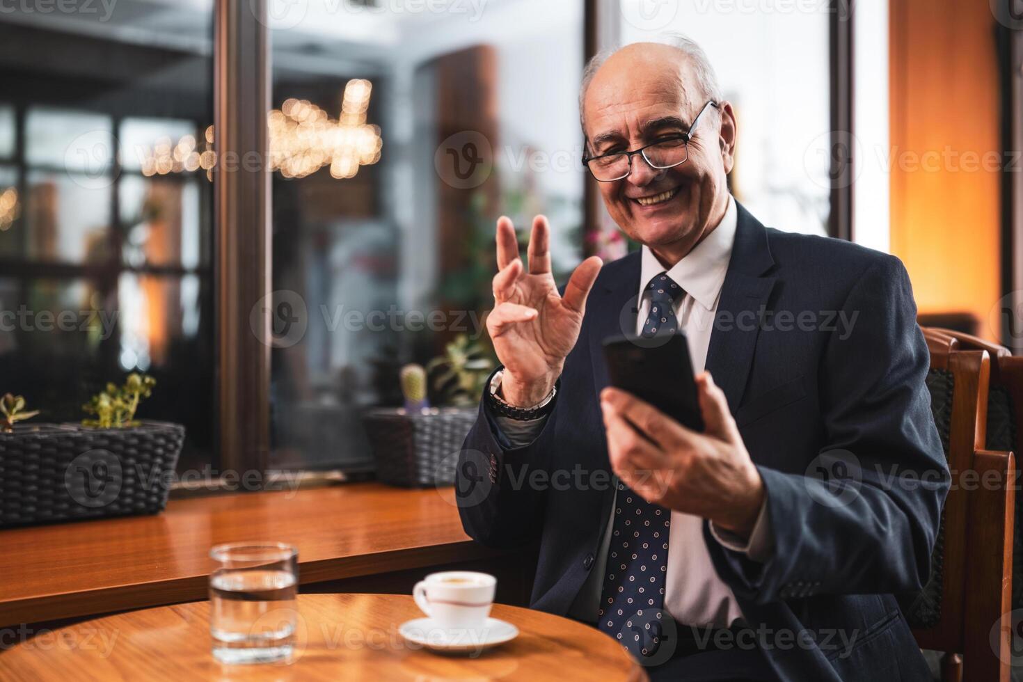 Senior businessman smiling and gesturing while holding mobile phone and sitting at the table in cafeteria. photo