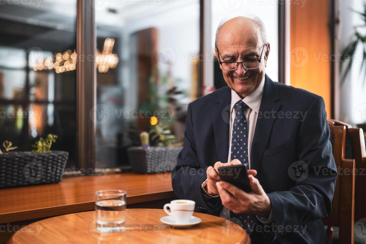 Smiling senior businessman with eyeglasses in elegant suit using mobile phone and drinking coffee while sitting in the cozy city cafe. photo
