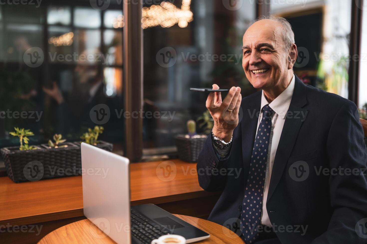 Confident older businessman in suit recording voice message via mobile phone and using laptop in cozy cafeteria. photo