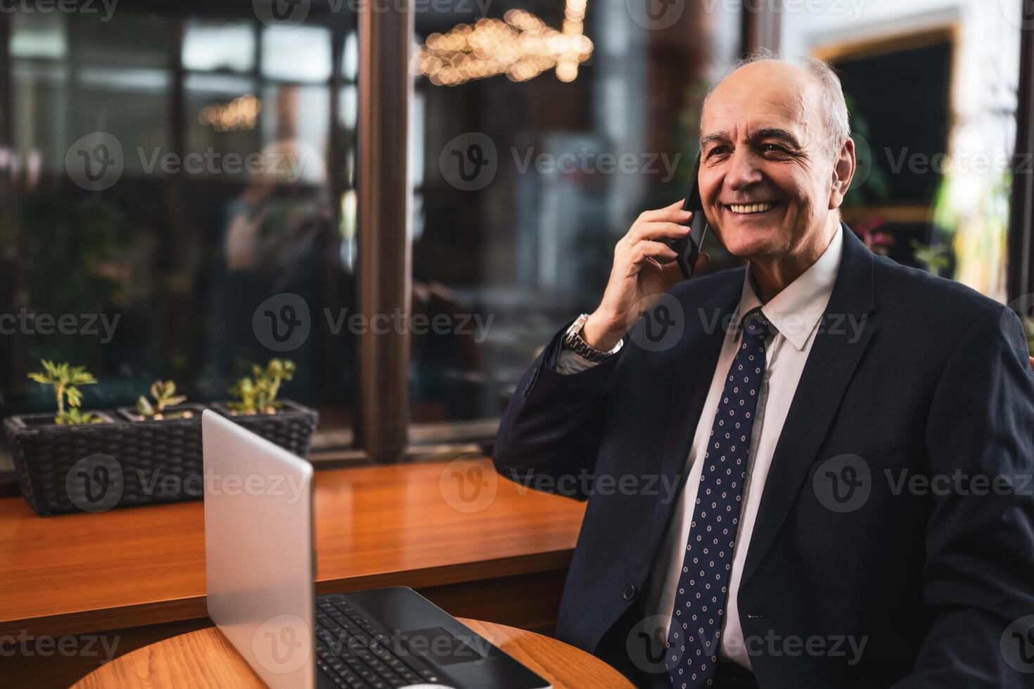 Senior businessman having a conversation on the phone and working laptop while sitting at the table in cafeteria. photo