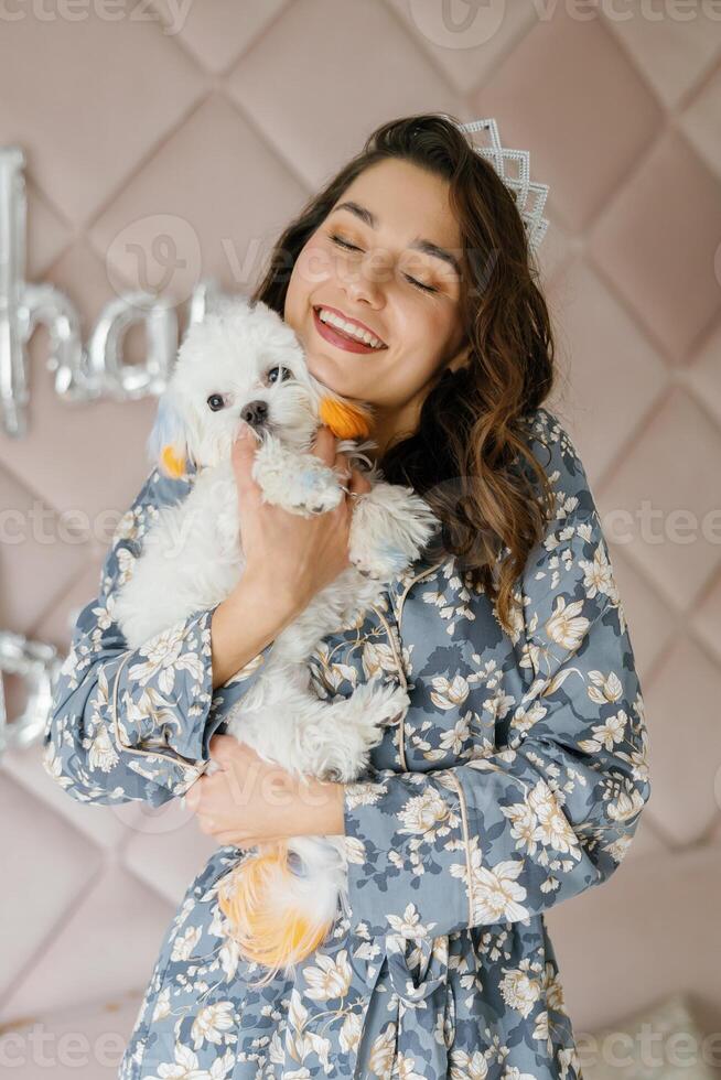 Smiling woman wearing crown holding adorable white dog with colorful fur photo