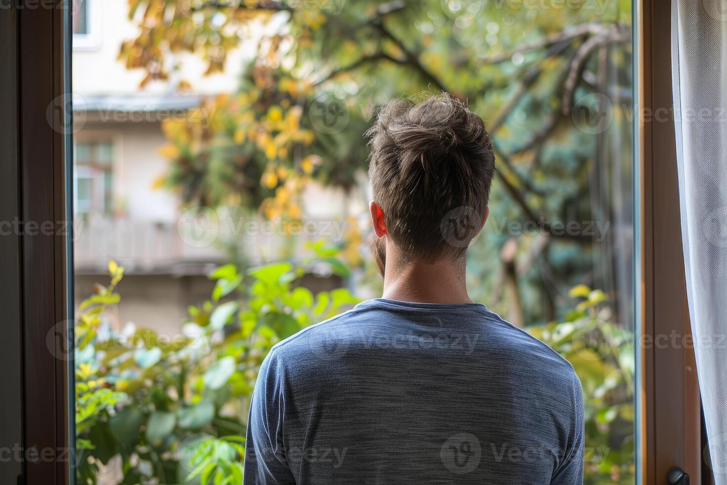 Man looking out of window at green trees and buildings in autumn photo