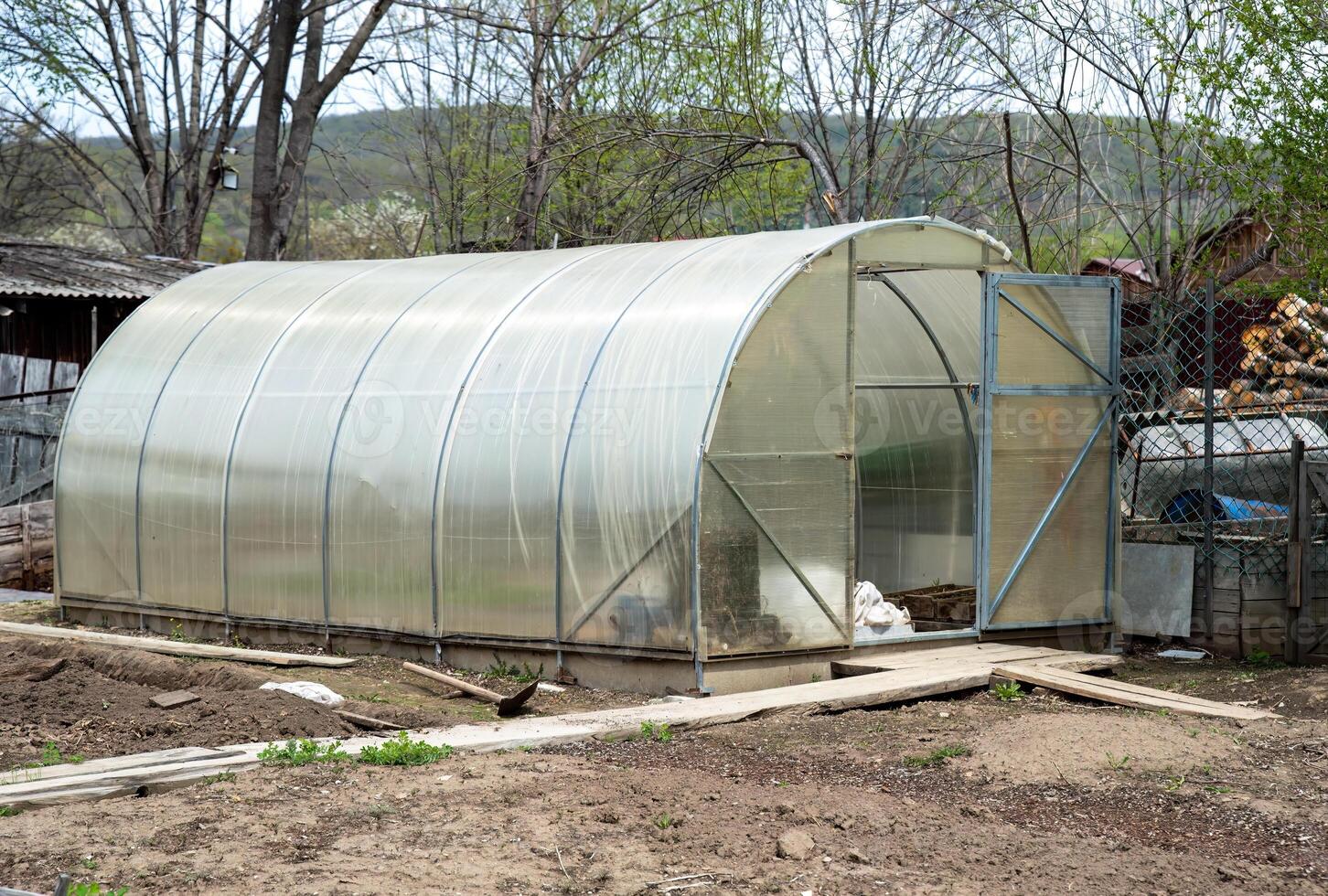 Open greenhouse in a backyard garden during spring. Transparent polycarbonate walls, surrounded by trees and gardening tools. Ideal for growing plants in rural settings photo