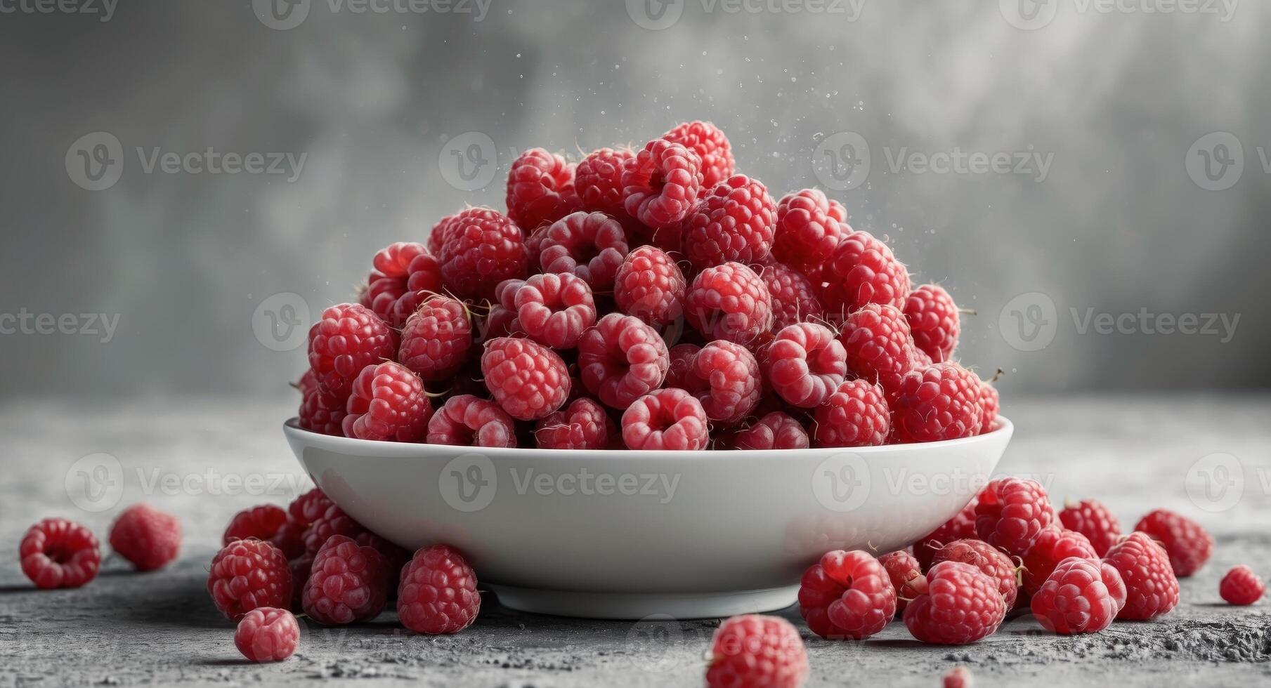 Fresh red raspberries overflowing in a white bowl on a textured gray concrete background with scattered berries and ample copy space. photo
