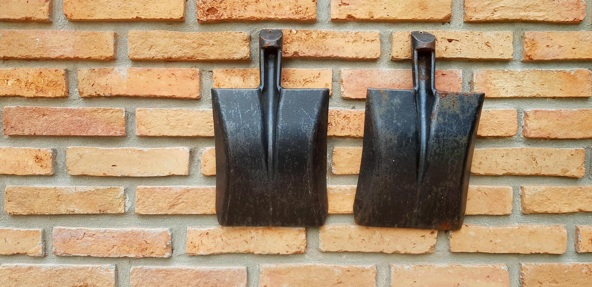 Two rusty black shovel, spade or hoe hanging on brick wall background with left copy space. Agriculture or Build tool, Vintage object and Decoration place concept. photo