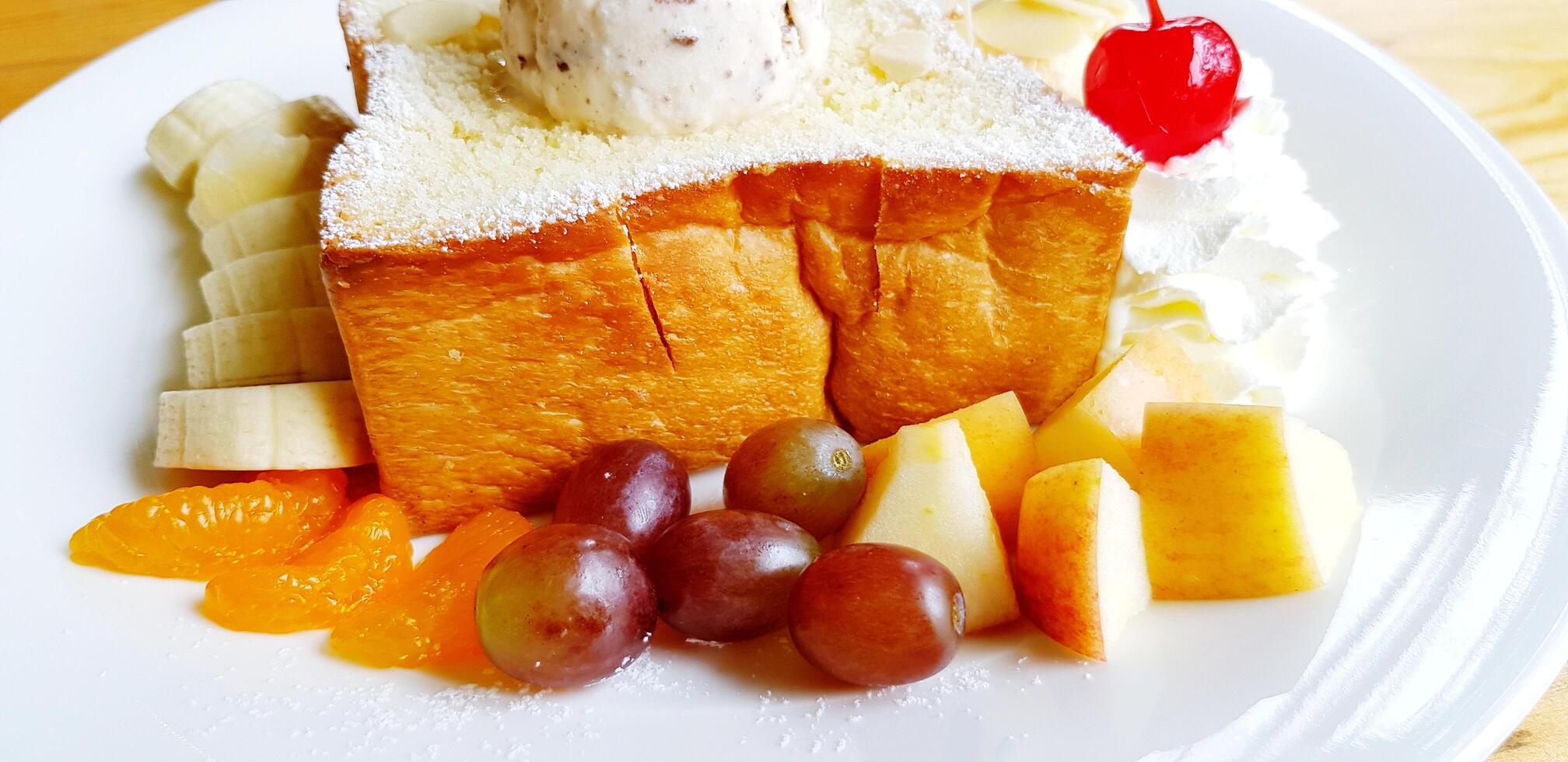 Close up sweet butter toast with top of chocolate chip ice cream sprinkle icing sugar, whipped cream with red cherry on top, vanilla ice cream and fresh fruit in white plate on wooden table. photo
