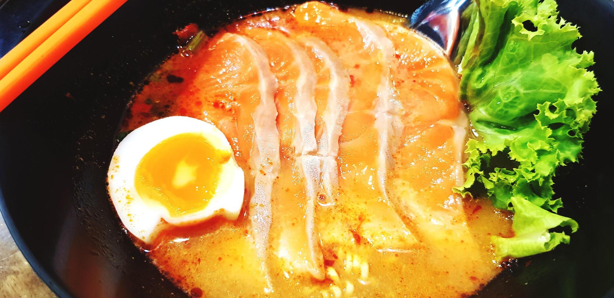 Close up hot spicy noodle soup with fresh salmon sliced, boiled yolk egg and green lettuce on black bowl at restaurant. photo