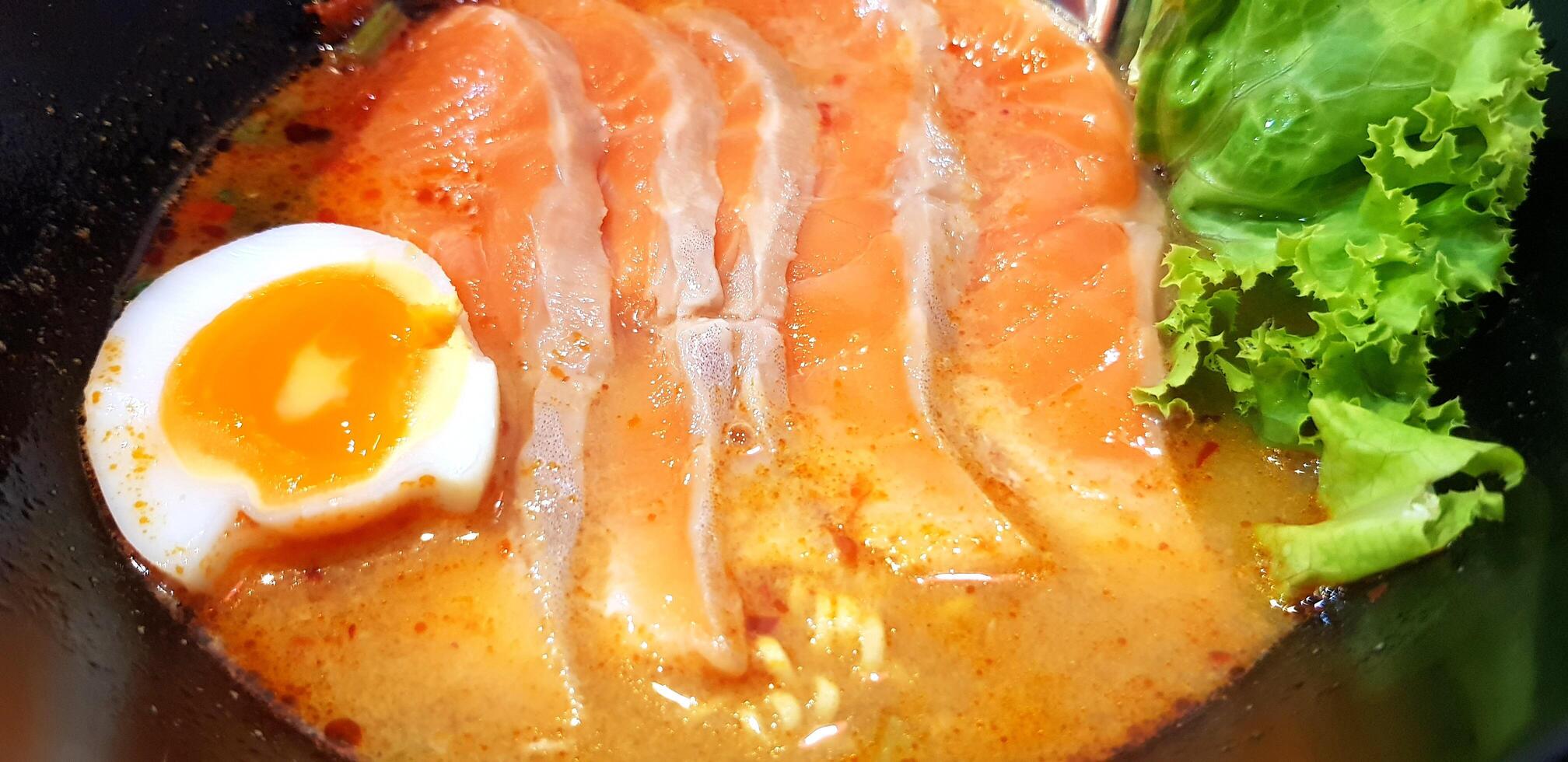 Close up hot spicy noodle soup with fresh salmon sliced, boiled yolk egg and green lettuce on black bowl at Japanese restaurant. photo