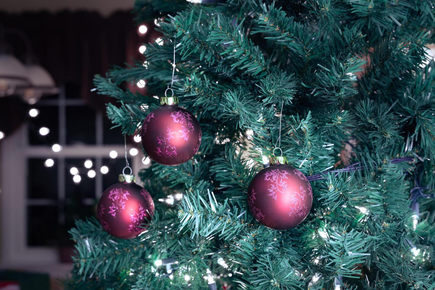 3 red Christmas tree balls hanging by a hook on a branch of an artificial pine tree decorated with lights. photo