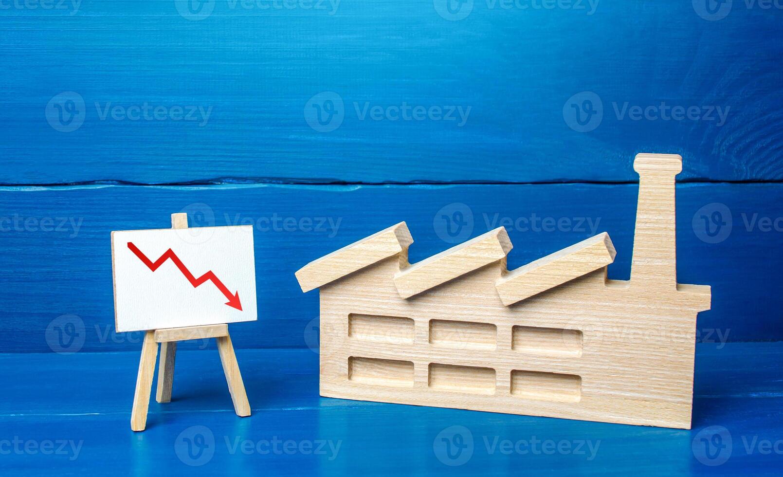 Figurine of industrial factory plant and easel with a red down arrow chart. Decreased production volumes, low income, energy resource consumption, emissions and pollution. Industry and economy fall. photo