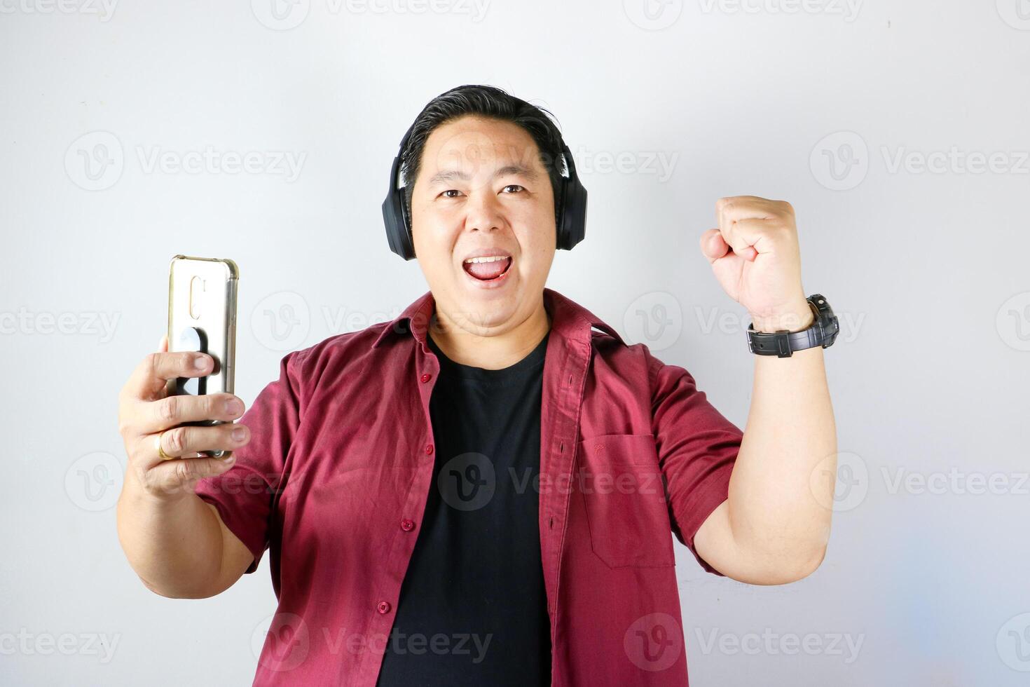 Cheerful handsome Asian Man holding his smartphone and wearing headphone while showing Wow expression and Yes gesture over isolated white background. photo
