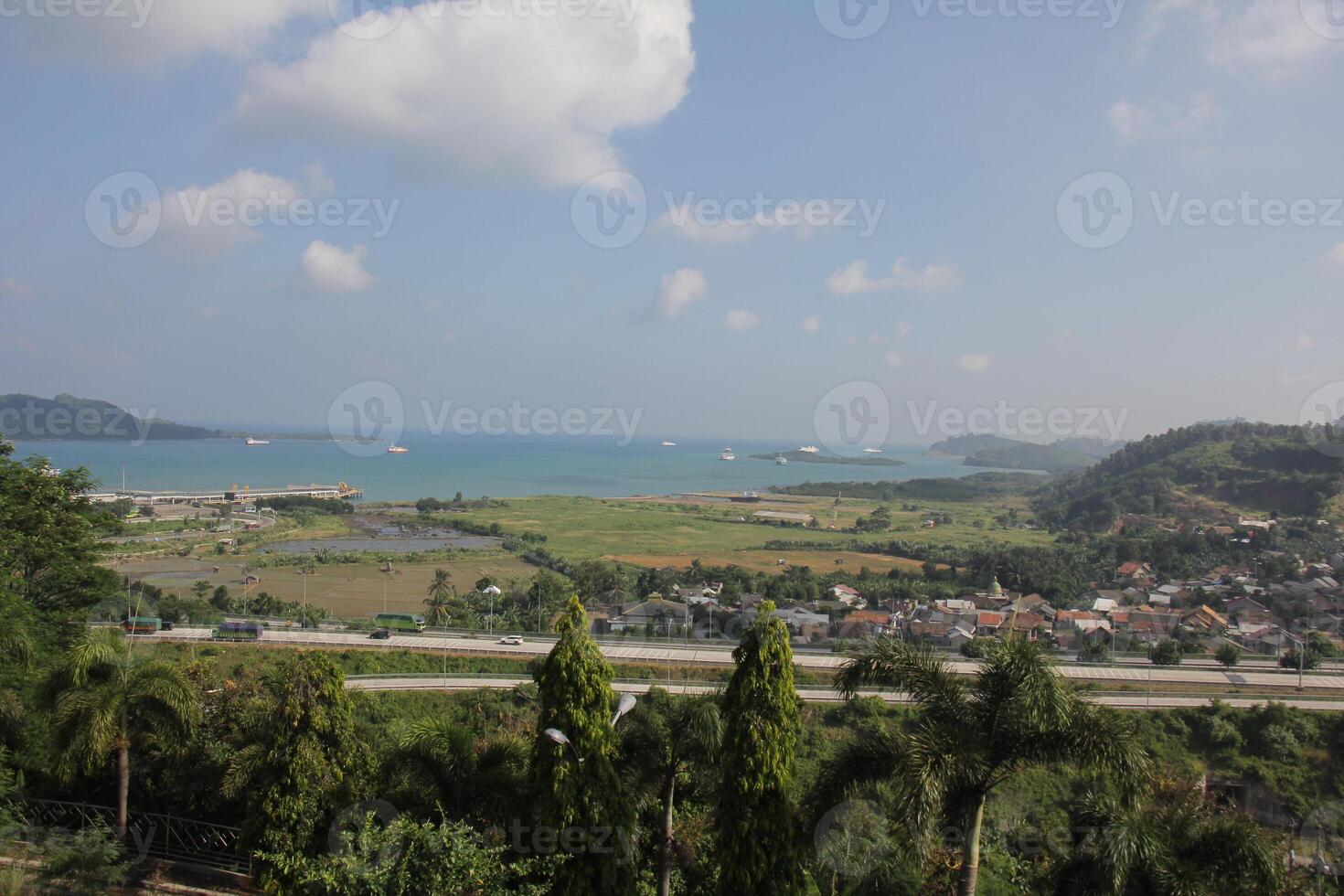 Panoramic view of a coastal town with a harbor, a highway and green fields under a cloudy sky photo