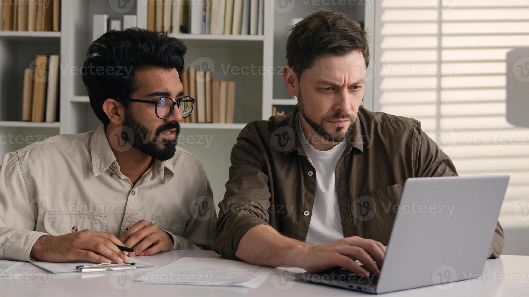 Two multiethnic colleagues businessmen business partners at office discuss financial data project documents paperwork Caucasian Indian men male coworkers look at laptop corporate teamwork discussion photo