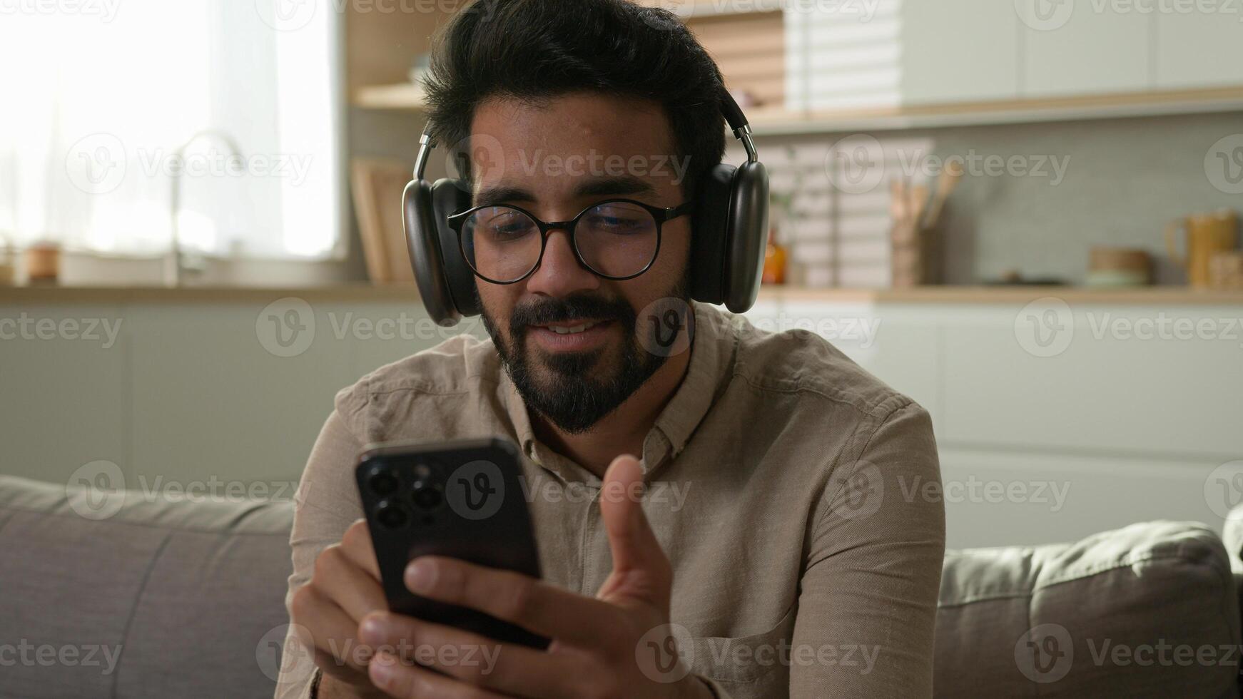 Carefree relaxed Arabian Indian man male ethnic guy in headphones listening music enjoy quality sound in wireless earphones disco musical track online audio service with mobile phone at home kitchen photo