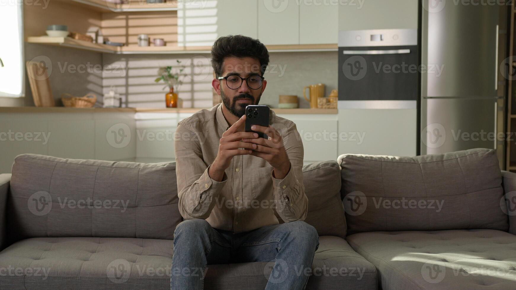 Arabian Indian ethnic man guy use mobile phone on couch browsing mobile internet at home kitchen take break confused read bad news on smartphone male freelancer scrolling cellphone app gadget addict photo