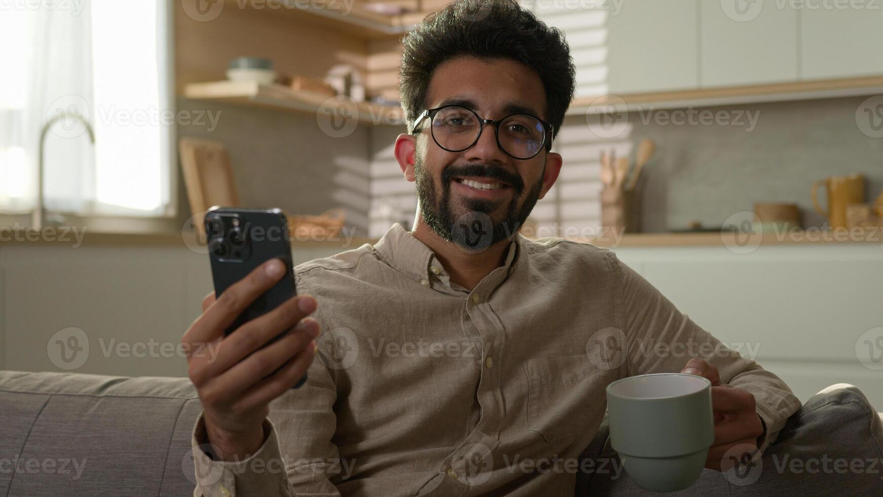 Relaxing Arabian man male ethnic guy on comfortable couch sofa using mobile phone drink coffee at kitchen Indian millennial freelancer working from home read news on smartphone looking at camera smile photo