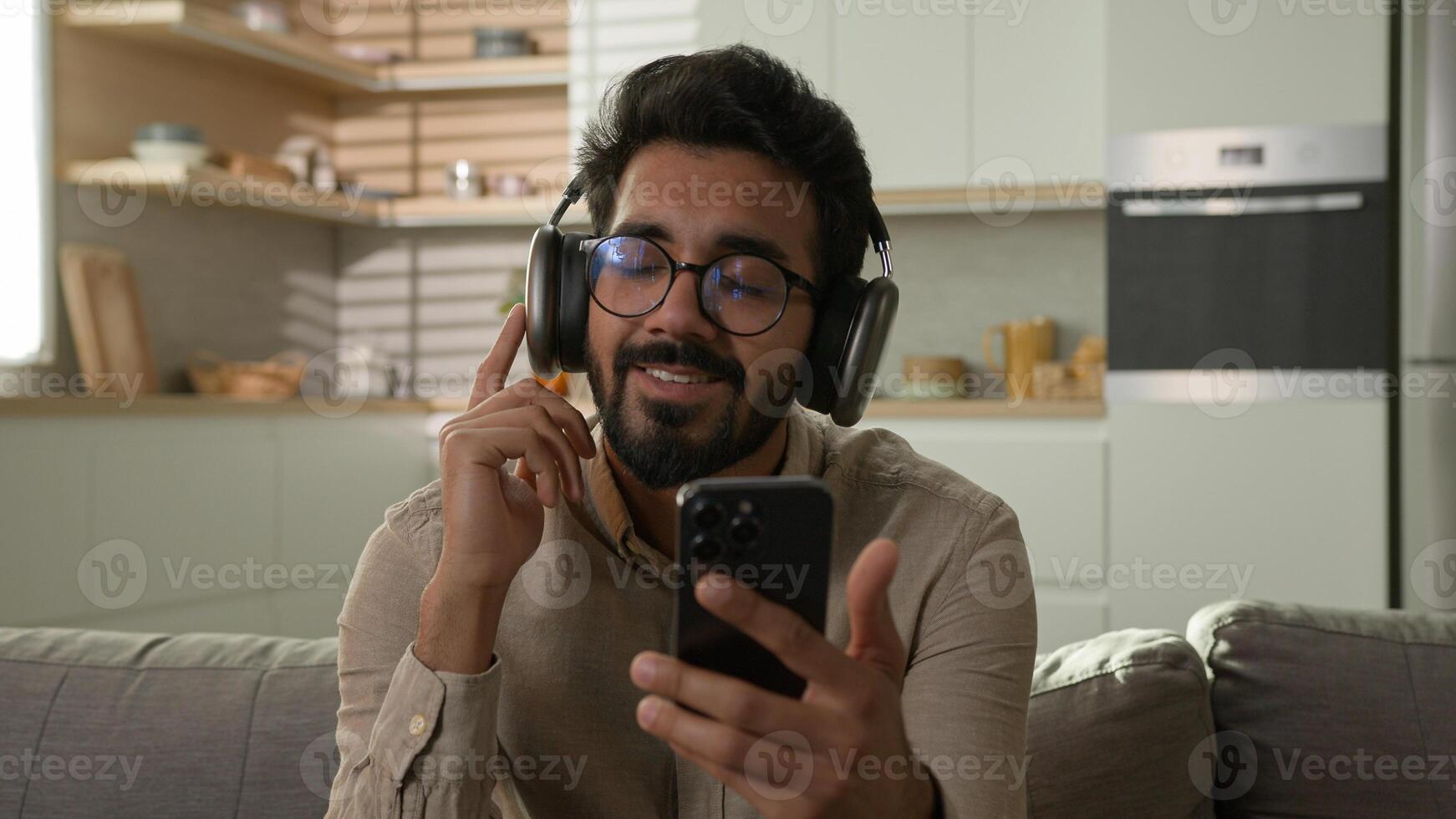 Relaxed happy man Indian Arabian guy male smiling choose audio song playlist on mobile phone smartphone listening music in wireless headphones earphones enjoy sound lazy weekend relax on couch at home photo