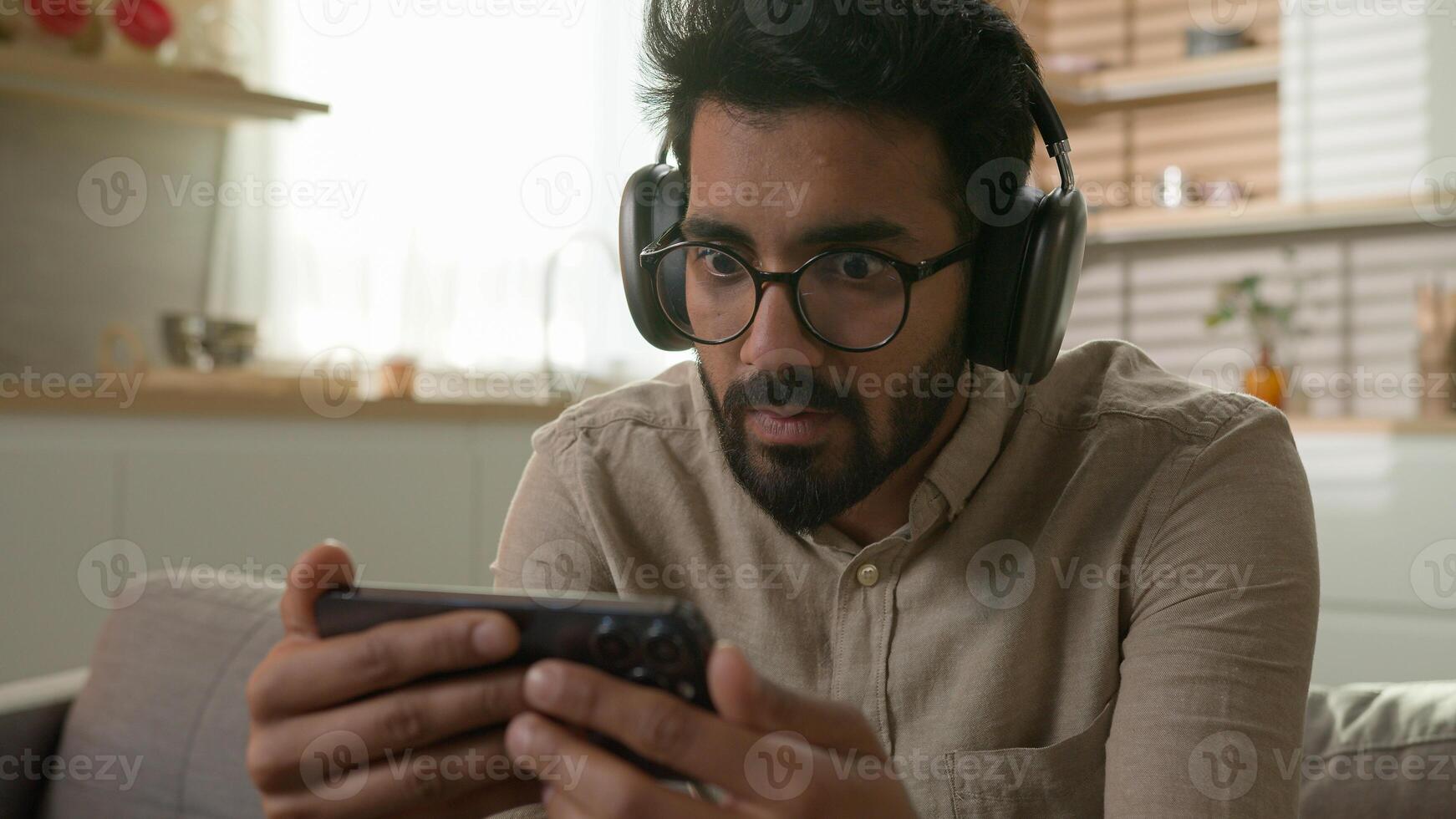 Disappointed Arabian Indian man guy watching sport game streaming on mobile phone upset by lose football match on couch in kitchen frustrated male in headphones looking at cellphone bad result loss photo