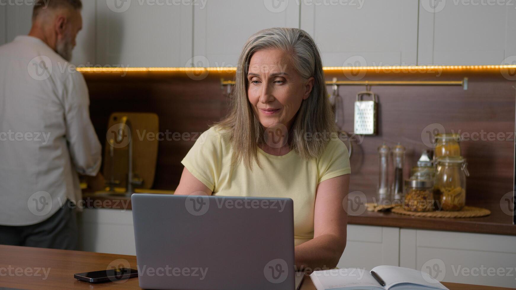 Mature 50s business woman wife online working with computer distance learning using laptop and mobile phone devices while senior man husband washing dishes in kitchen middle aged family couple at home photo