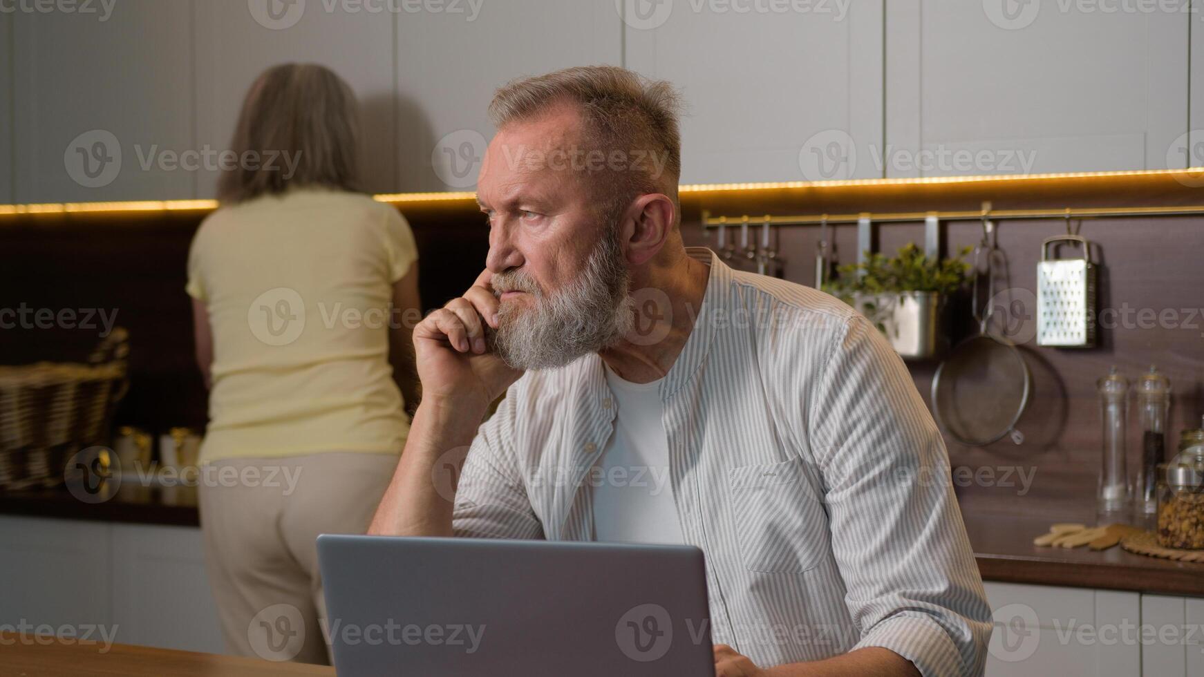 Middle aged retired 50s man husband gray-haired businessman thinking working distant with computer notebook thoughtful think problem solution laptop work at home while woman wife cooking in kitchen photo