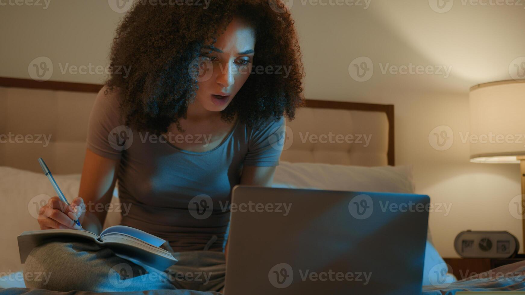 African American woman student high school learner pupil girl studying on bed use laptop watch lecture online prepare for exam night homework write notes in book learn subject e-learning at home photo