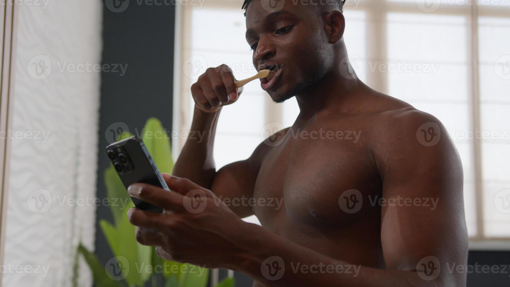 African American bare multiracial man guy brushing teeth with toothbrush morning routine dental hygiene brush with toothpaste oral tooth care in bathroom using mobile phone read media on smartphone photo