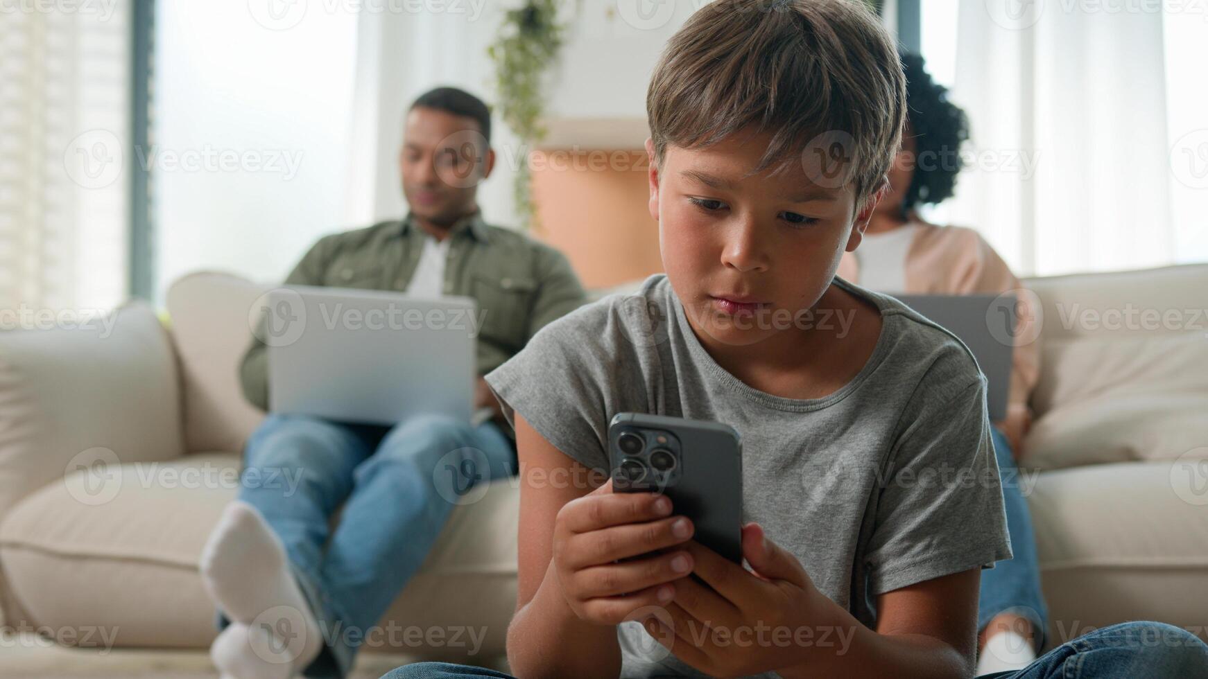 Gadget addicted family with devices at home multiracial mom and dad using laptops addict work ignoring sad child little boy son bored kid with phone smartphone on floor lack parents attention custody photo