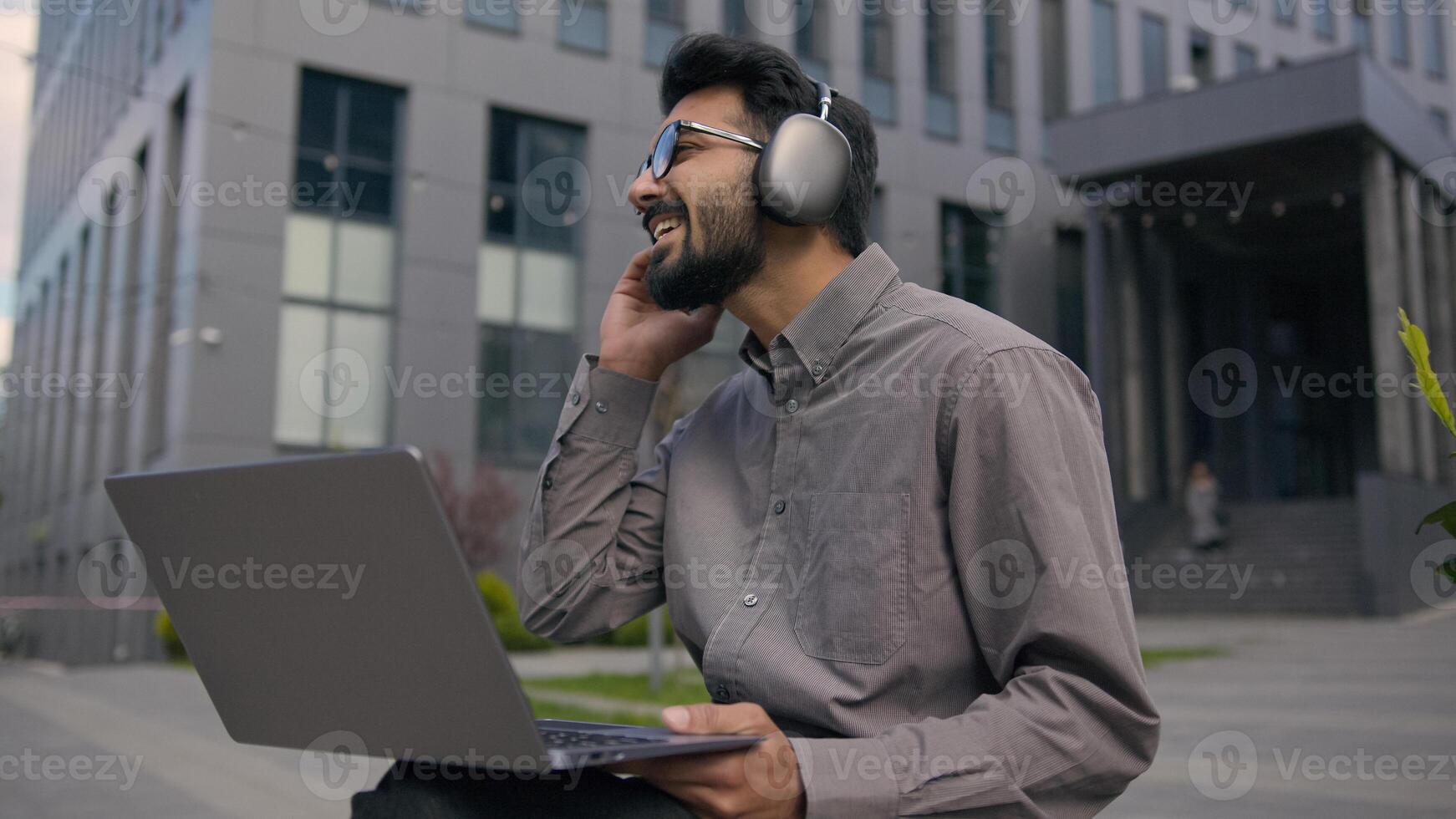 Arabian Indian man listen music educational webinar in headphones outdoors male businessman freelancer work with laptop develop business project online studying smiling guy student e-learning in city photo