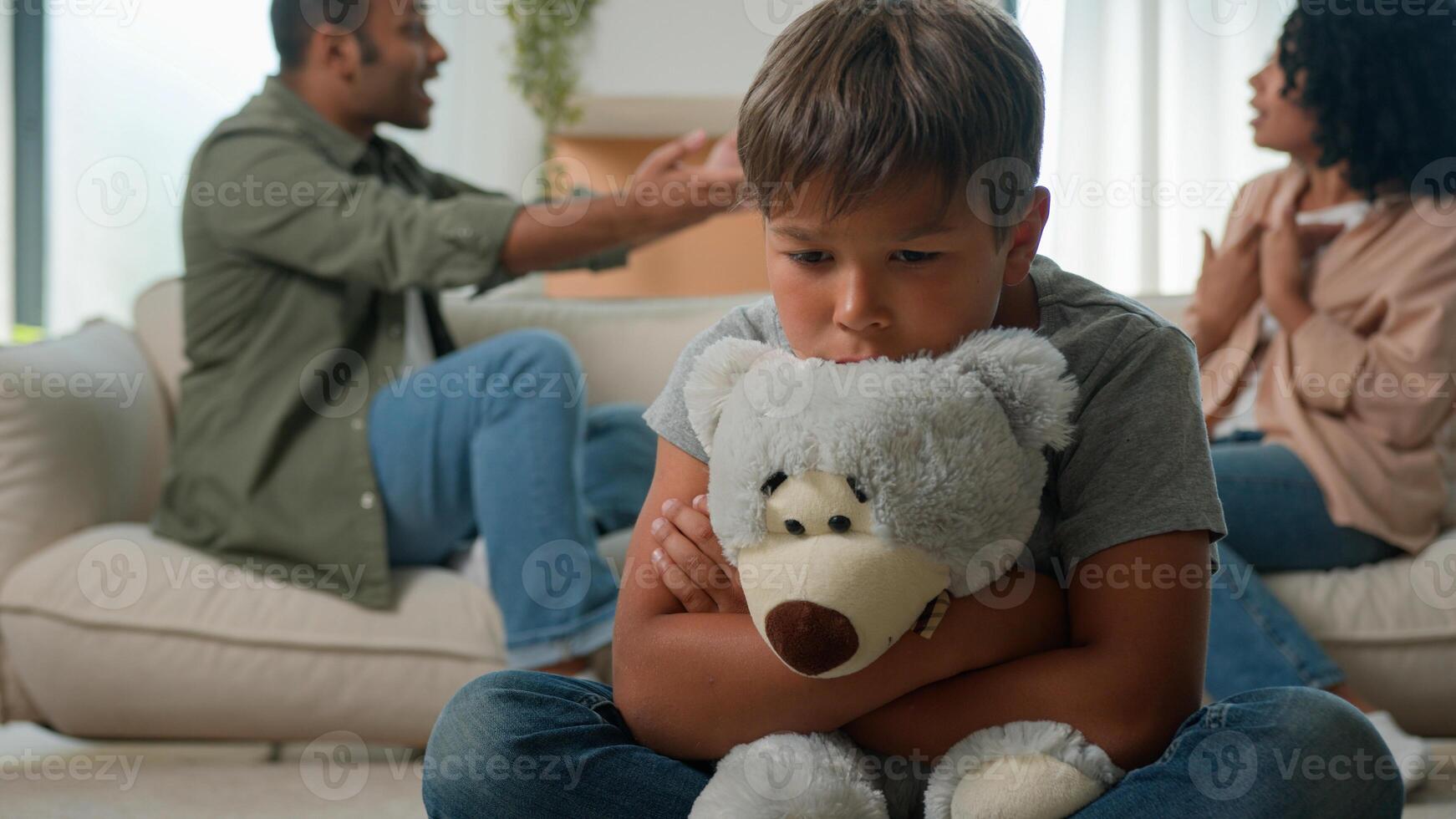 Upset Caucasian little boy preschool son hug teddy toy while African American woman mother and man father in anger screaming arguing background sad kid suffer from family quarrel conflict child trauma photo