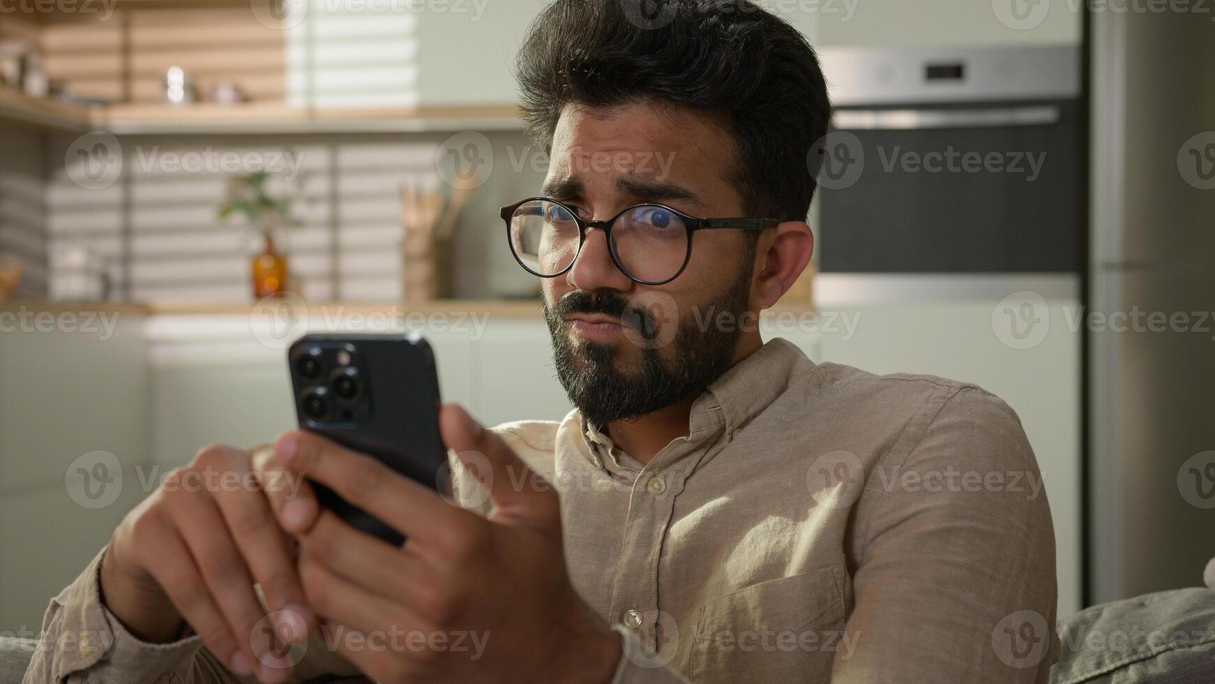 Arabian Indian ethnic man guy use mobile phone on couch browsing mobile internet at home kitchen take break confused read bad news on smartphone male freelancer scrolling cellphone app gadget addict photo