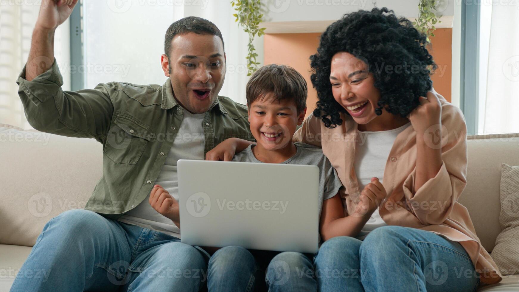 Happy family African American parents mum dad and Caucasian little boy kid child teenager son use laptop playing game together watch fun cartoons win bet online victory celebrate on sofa at home photo