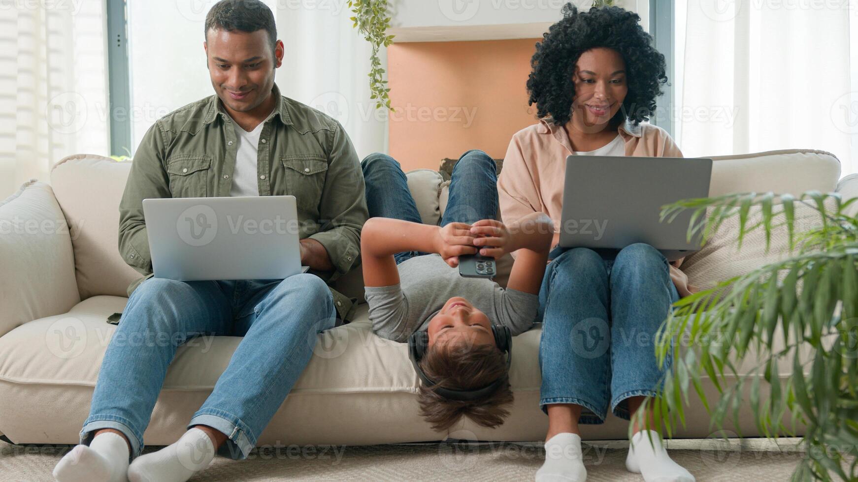Addicted family use gadgets devices little kid child son boy lying upside down on sofa play game on mobile phone smiling African American parents working laptops different gen using modern tech photo