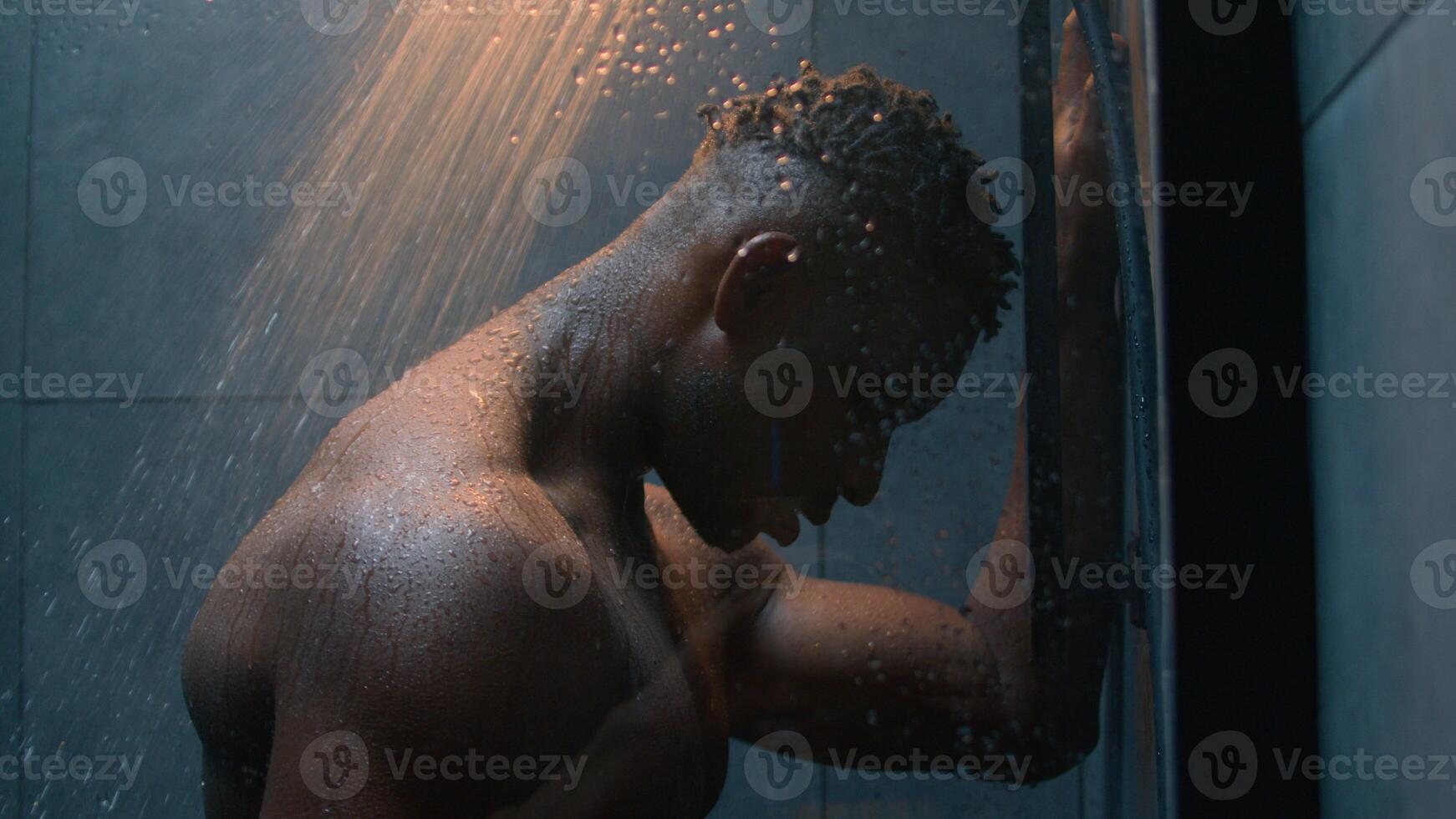 African American naked muscular man washing in dark bathroom take shower under falling water drops rain wet tired exhausted guy showering after workout refresh body daily hygiene male bodycare in bath photo
