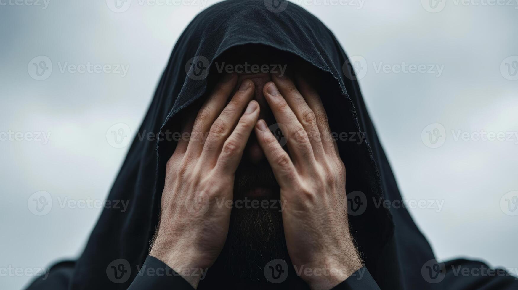 Individual in dark hood covering face with hands in an outdoor setting under cloudy sky photo