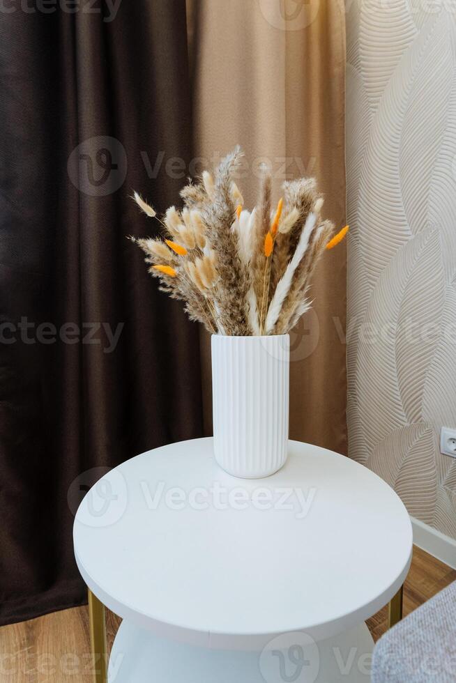 A stunningly crafted vase filled with an assortment of dried flowers in a range of vibrant colors is gracefully positioned on a sleek white table, enhancing the charming decor of the room photo
