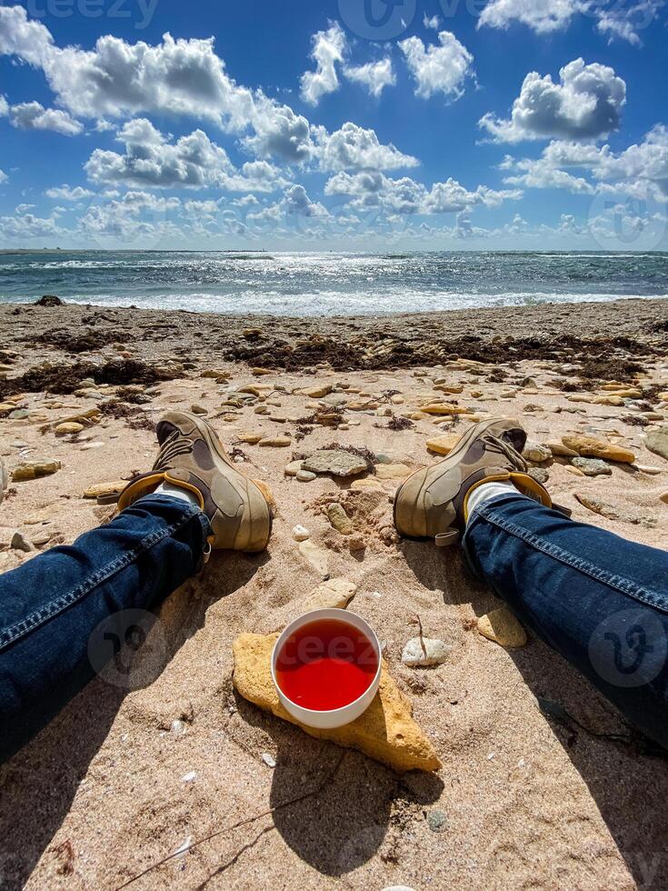 A person is relaxing while laying on the warm beach, holding a cup filled with a vibrant red liquid that catches the sunlight beautifully, enjoying the serene coastal view photo