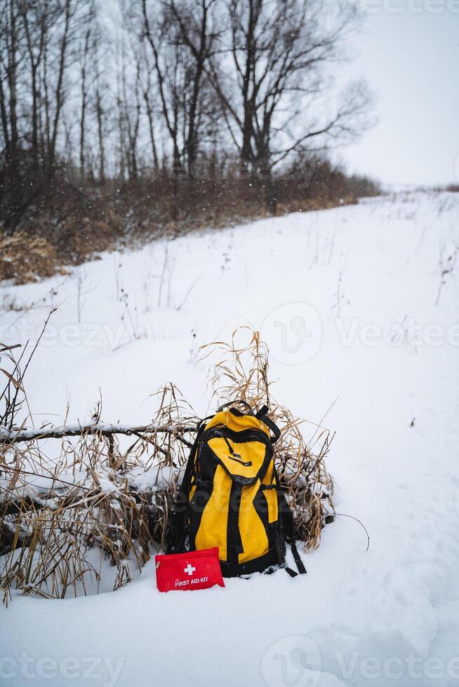 A vibrant yellow backpack is resting in the snow, which has blanketed the ground, while a bright red first aid kit is placed beside it, waiting to be used in case of emergencies photo