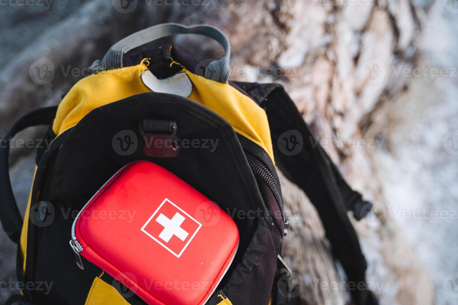 A bright red first aid kit is positioned neatly on top of a vibrant yellow backpack, making it an easily recognizable and essential piece of equipment for any outdoor journey or adventure photo