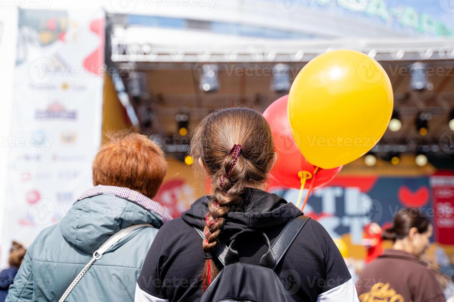 A woman, wearing a backpack, is joyfully holding a bright yellow balloon as she stands in front of a stage, surrounded by people at a lively event, creating a fun atmosphere photo