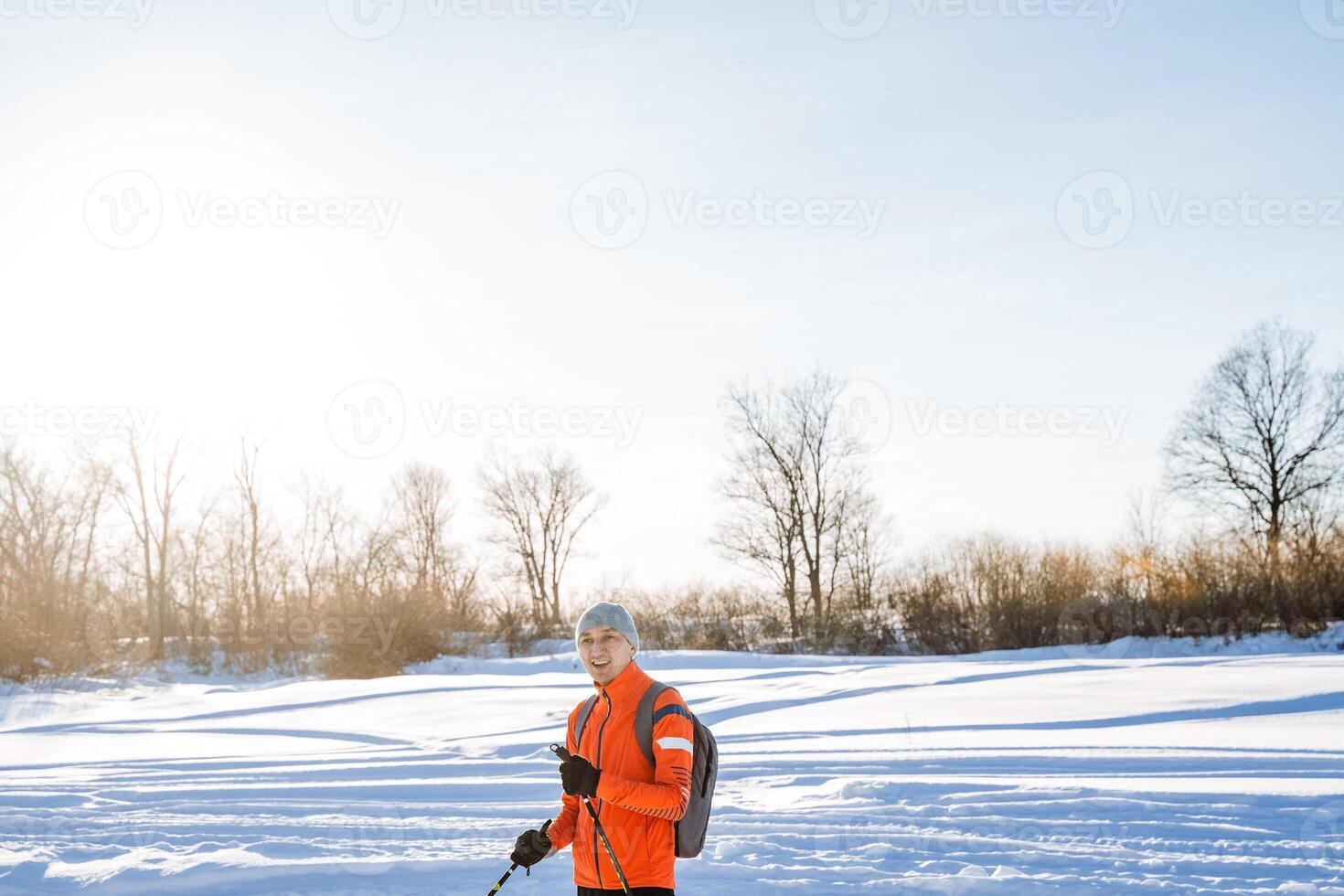 Man in orange tracksuit standing on the background of snow and winter forest, outdoor workout in nature, skier with poles. photo