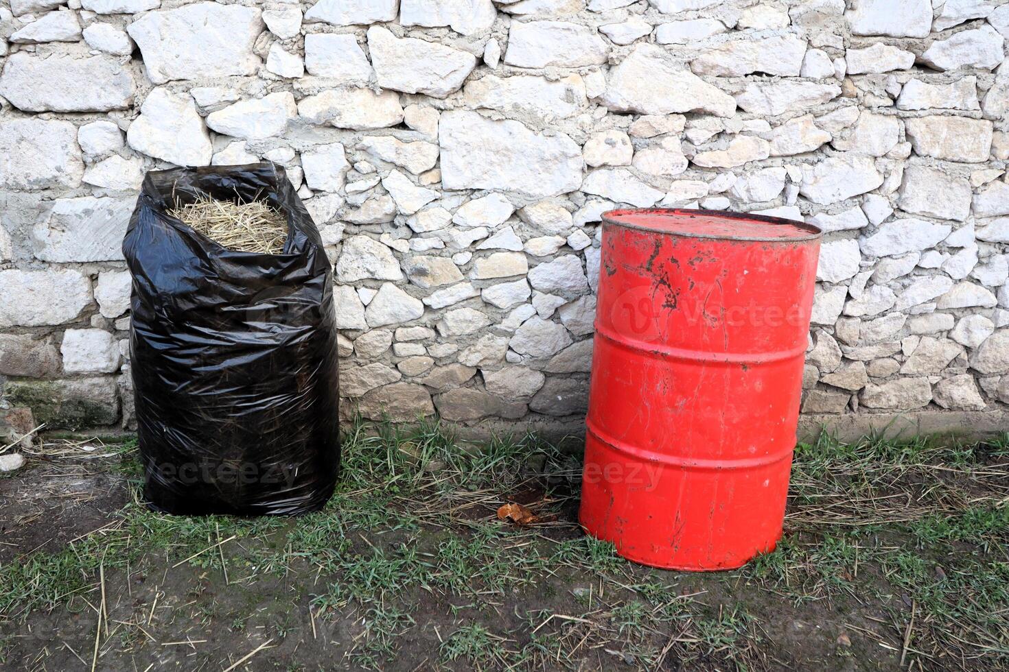 The waste disposal setup includes a black bag and a red barrel placed against a stone wall photo