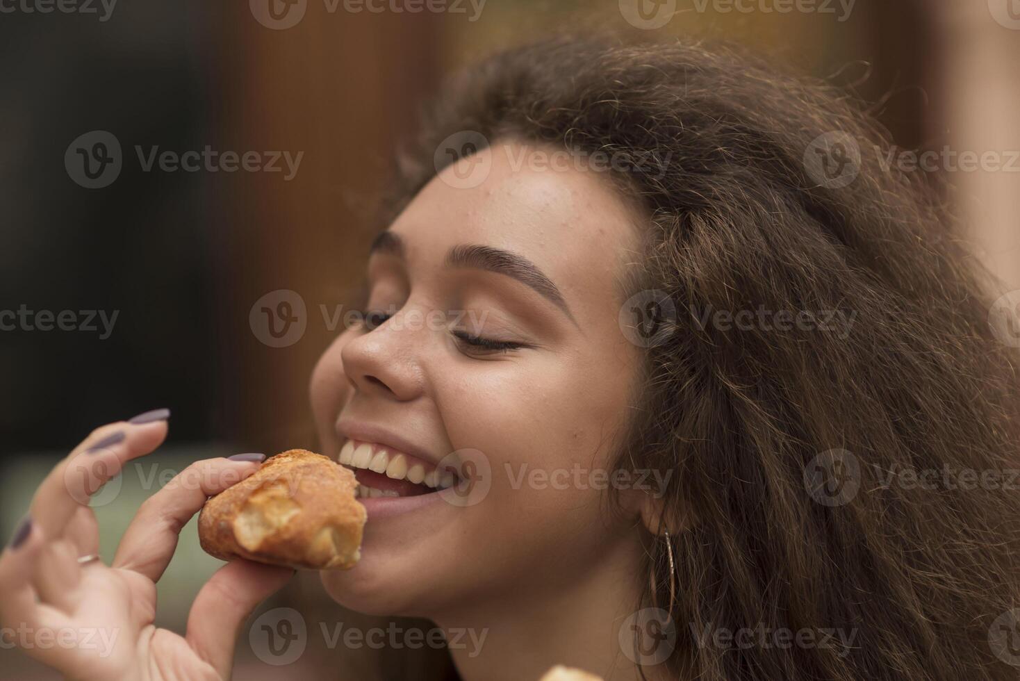 A young girl eats a baguette with appetite. She has a piece of bread in her hands and is going to eat it. She has a pleasant smile. The girl has wavy fluffy hair. A close-up of her face. photo
