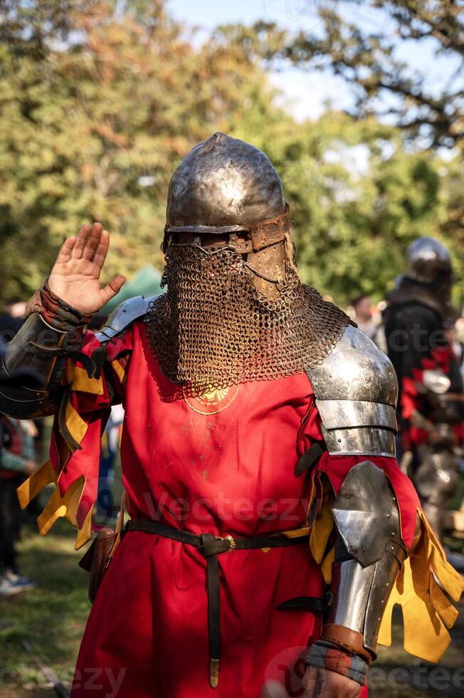 a man in medieval armor. a man dressed as a knight from the Middle Ages. On him is a helmet and iron armor. he is wearing a red shirt. the man waves his hand, the photo was taken outdoors