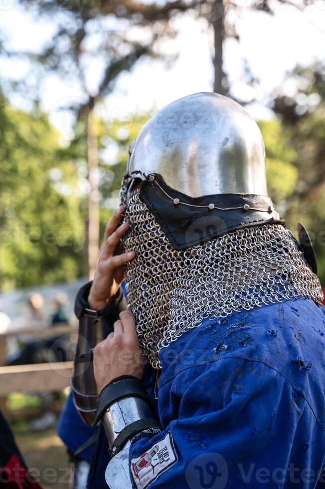 a man in medieval armor. a man dressed as a knight from the Middle Ages. On him is a helmet and iron armor. he wears a blue shirt. The man is fixing his helmet, the photo was taken outdoors