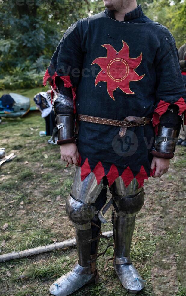 a man in the costume of a medieval knight. a man stands in iron armor. his face is not visible. a red sun is embroidered on his medieval shirt. photo taken vertically